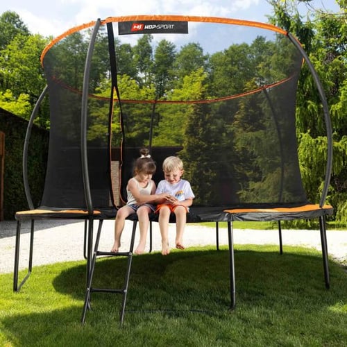 twee kinderen zitten op de mat van een ronde Hop-Sport trampoline met oranje veiligheidsnet en ladder tijdens trampolinespringen, kijkend naar een tablet, tuin op de achtergrond