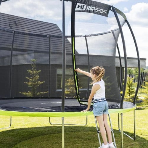 meisje klimt via de ladder op een ronde Hop-Sport trampoline met groene mat en veiligheidsnet tijdens trampoline trucs, huis op de achtergrond