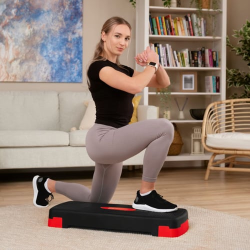 vrouw wil afvallen met step-aerobics en doet een lunge-oefening met één knie bij een zwart-rode step in de woonkamer
