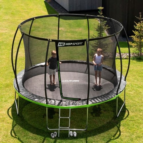 twee kinderen staan op een ronde Hop-Sport trampoline met groene mat en veiligheidsnet tijdens trampoline trucs, tuin op de achtergrond