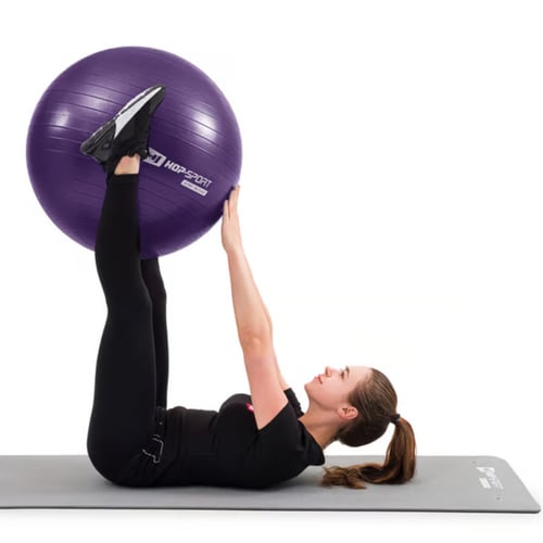 vrouw ligt op haar rug op een mat en houdt haar voeten op een paarse Hop-Sport fitnessbal boven zich, met haar handen ter ondersteuning